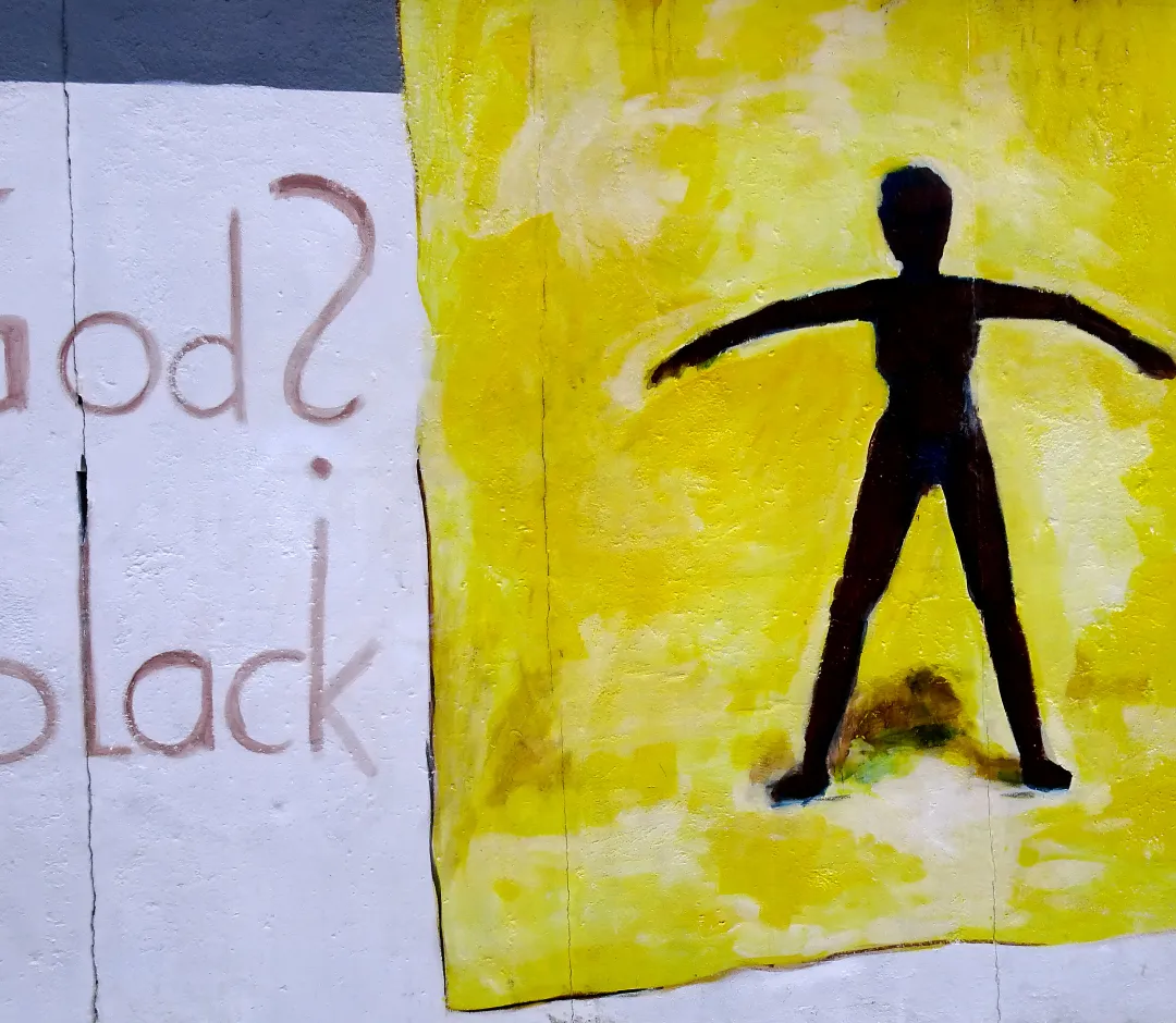 Ausschnitt eines Wandgemäldes der Berliner East Side Gallery: Links steht der Schriftzug God? black, rechts daneben eine dunkle menschliche Silhouette mit ausgebreiteten Armen vor einem leuchtend gelben, rechteckigen Hintergrund.