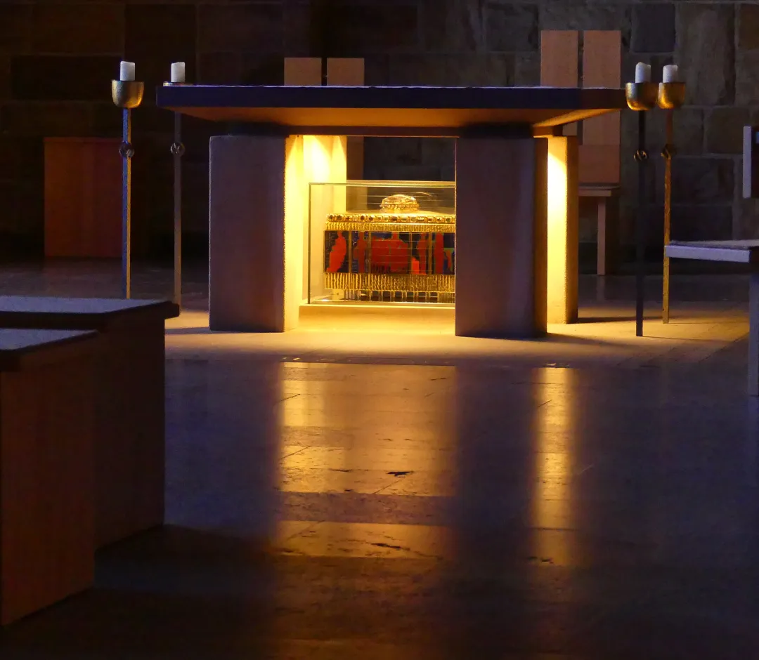 Im Zentrum eines dunklen Kirchenraums steht ein beleuchteter Altar aus Stein, unter dessen Platte ein farbenfroh gestalteter Schrein hinter Glas in warmem Licht erstrahlt. Der polierte Steinboden im Vordergrund reflektiert das Licht, während die umliegenden Holzstühle und Kirchenbänke weitgehend im Schatten liegen.