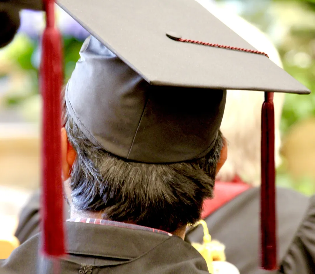 Nahaufnahme von hinten auf eine schwarze Graduiertenkappe mit roter Quaste, getragen von einer Person bei einer Examensfeier vor einem unscharfen Hintergrund.