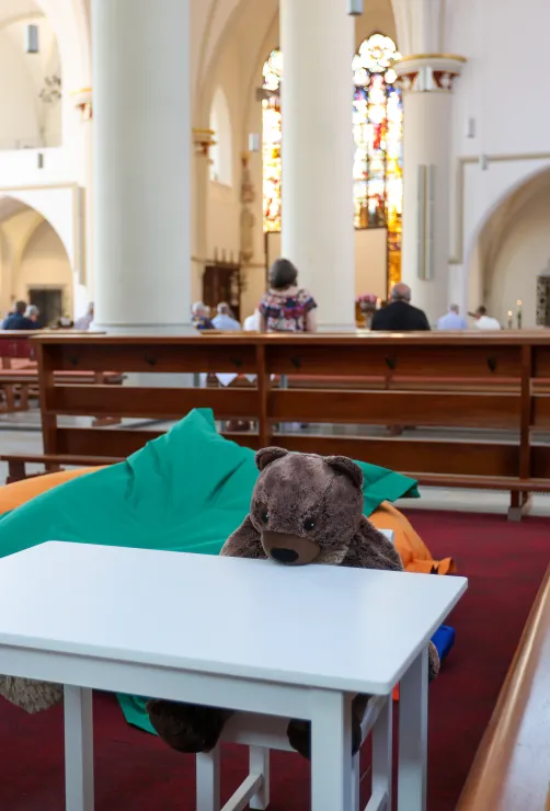 Kinderecke in einer Kirche: Plüschtiere sitzen um einen weißen Tisch. Im Bildhintergrund feiern Erwachsene den Gottesdienst.