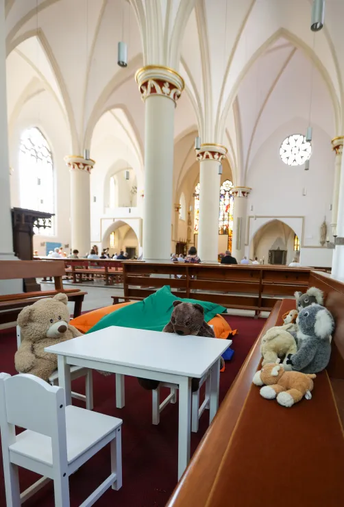 Kinderecke in einer Kirche: Plüschtiere sitzen um einen weißen Tisch. Im Bildhintergrund feiern Erwachsene den Gottesdienst.