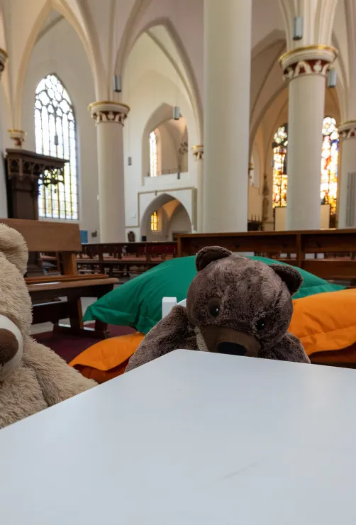 Ein Plüschbär und weitere Plüschtiere sitzen im Bildvordergrund einer hellen Kirche. Neben ihm liegen bunte Kissen am Boden, im unscharfen Hintergrund sind Kirchenbänke, Säulen und gotische Bögen zu sehen.