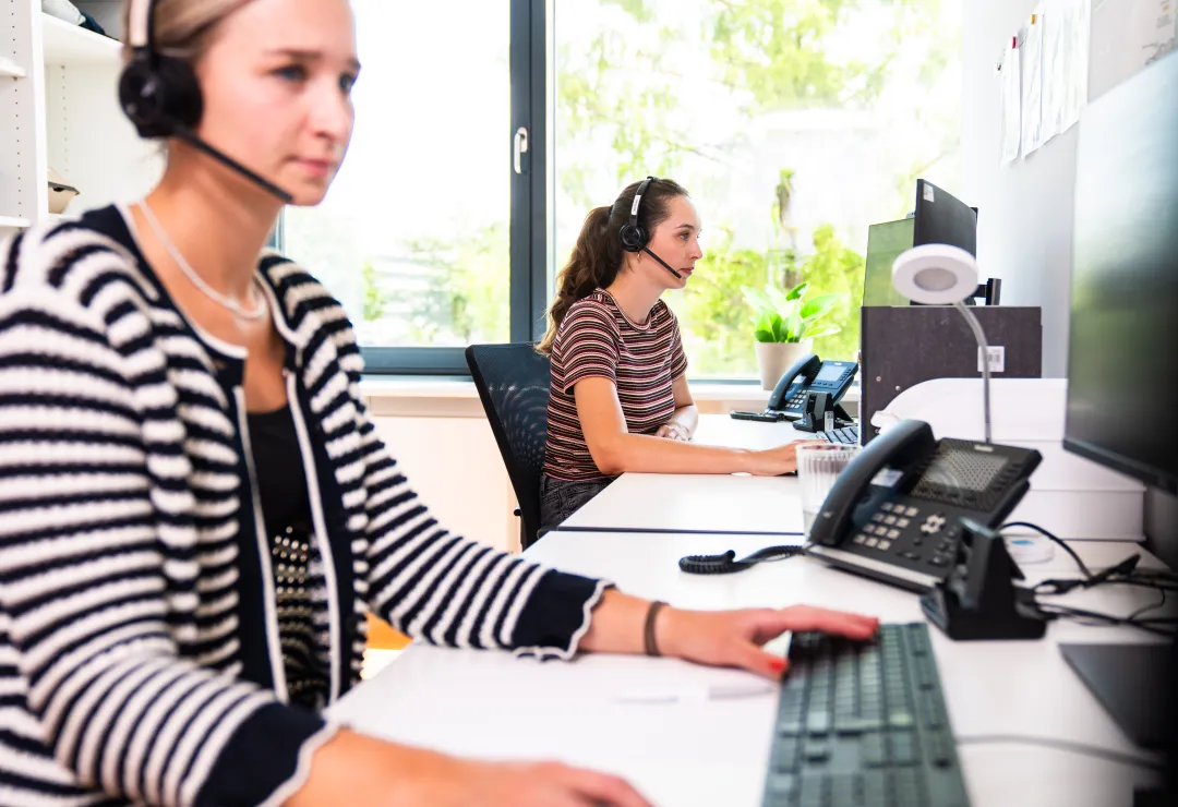 Zwei Frauen mit Headsets arbeiten an Computern in einem modernen Büro.