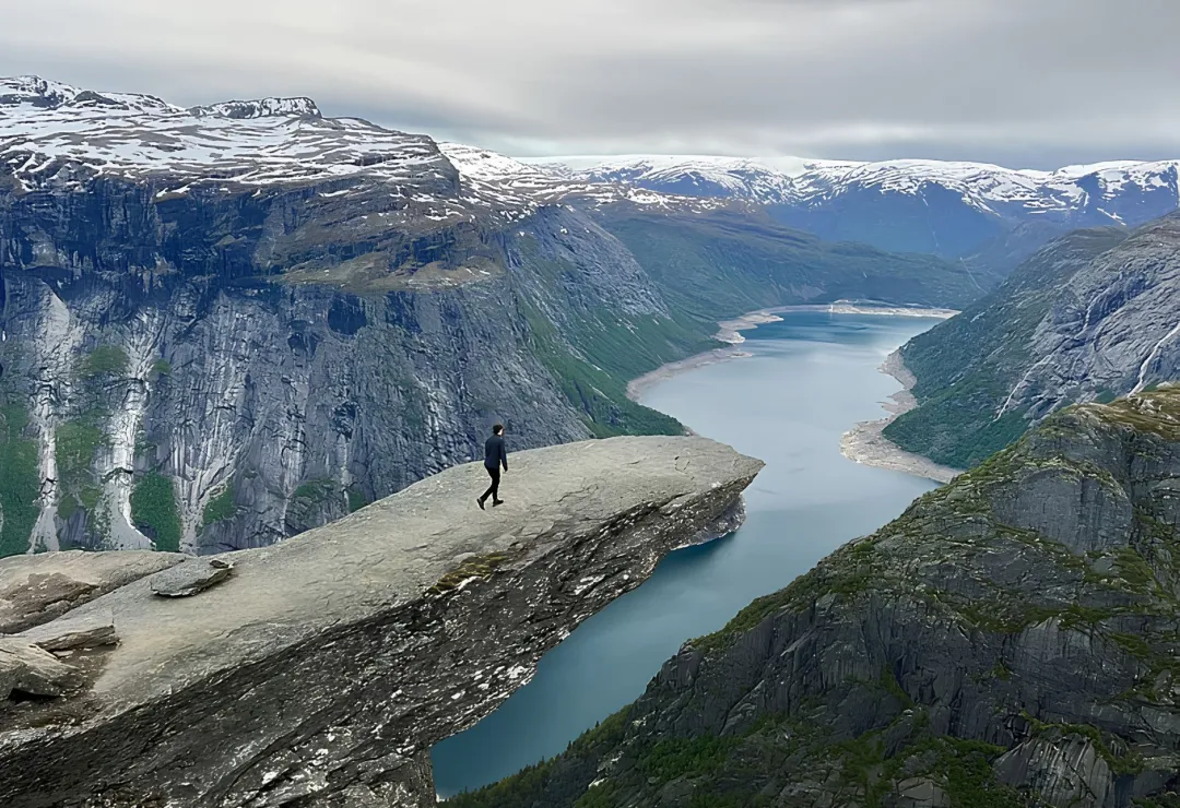 Person steht auf Felsvorsprung über Fjord mit Bergen und bewölktem Himmel