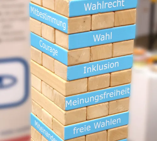 Ein Turm aus Holzbausteinen, auf denen teils Begriffe stehen, die für eine Demokratie wesentlich sind.