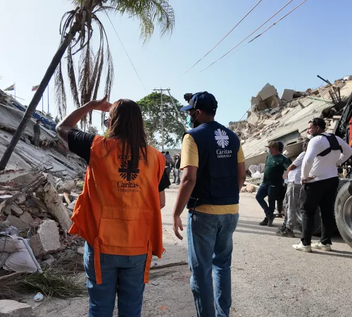 Zwei Mitarbeiter von Caritas International stehen auf de Straße vor einem von einem Erdbeben zerstörten Haus.