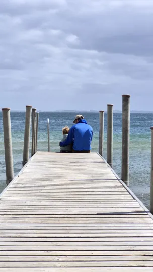 Ein Mann mit Jacke und Schildmütze sitzt zusammen mit einem kleinen Kind am Ende eines hölzernen Stegs. Der Mann hält den Arm schützend um das Kind. Beide schauen hinaus auf das ruhige, leicht wogende Meer und den bewölkten Himmel.