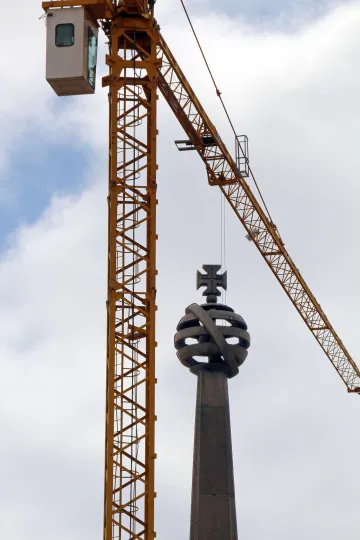 Ein Kran, unter dessen Ausleger sich ein Kirchturm erstreckt. Im Hintergrund ist ein bewölkter Himmel zu sehen.