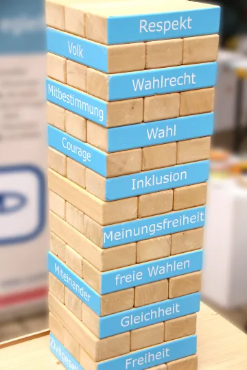 Ein Turm aus Holzbausteinen, auf denen teils Begriffe stehen, die für eine Demokratie wesentlich sind.