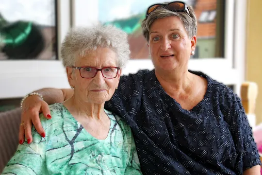 Eine alte und eine jüngere Frau sitzen nebeneinander auf einer Bank. Die Jüngere hält die Ätere im Arm.