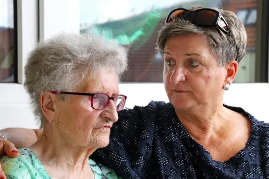 Eine alte und eine jüngere Frau sitzen nebeneinander auf einer Bank. Die Jüngere hält die Ätere im Arm.