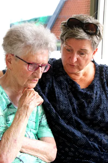 Eine alte und eine jüngere Frau sitzen nebeneinander auf einer Bank. Die Jüngere hält die Ätere im Arm.