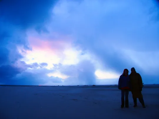 Zwei Personen stehen bei Sonnenuntergang am Strand, Himmel leuchtet in Blau- und Rosatönen.