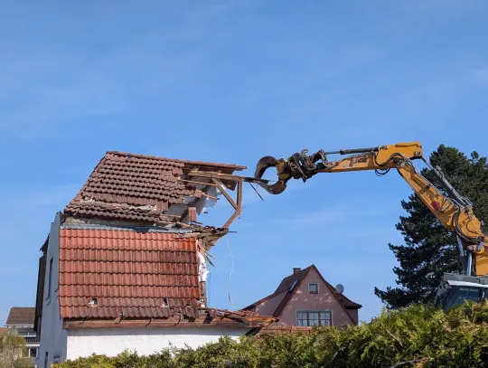 Ein gelber Bagger reißt mit einem Greifarm das Dach eines alten Wohnhauses mit roten Ziegeln ein. Im Hintergrund ist ein blauer Himmel und ein benachbartes Haus sichtbar, im Vordergrund eine grüne Hecke.
