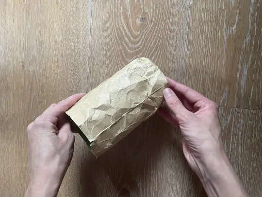 Zwei Hände halten einen Tetrapak aus braunem Papier über einem Holzboden.
