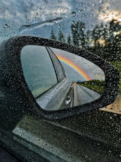 Blick durch eine von Regentropfen benetzte Autoscheibe auf den Außenspiegel, in dessen Reflexion ein farbenfroher Regenbogen über einer nassen Straße und dunklen Wolken zu sehen ist.