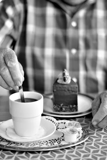 Schwarz-Weiß-Nahaufnahme der faltigen Hände einer älteren Person in einem karierten Hemd, die an einem Tisch in einem Café sitzt. Eine Hand rührt mit einem Löffel in einer weißen Kaffeetasse, die auf einem silbernen Tablett mit Spitzendecke steht. Im Hintergrund ist ein Stück Schokoladentorte auf einem Teller in Unschärfe zu sehen.