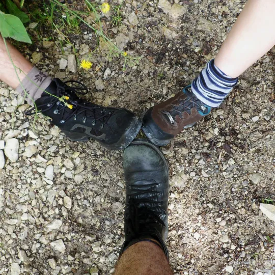 Drei Füße in Wanderschuhen, die im Kreis zueinander zeigen. Sie stehen auf einem Kiesweg. Die Schuhe und Socken sind alle verschieden und deuten auf unterschiedliche Personen hin, die gemeinsam unterwegs sind.