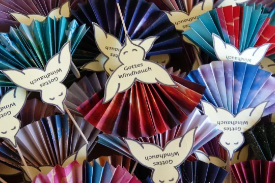 Gebastelte Taubenfiguren aus Papier. Auf dem hellgelben Körper des Vogels steht der Text "Gottes Windhauch", die Flügel sind als farbige Papierfächer gestaltet.