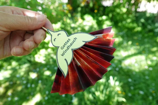 Eine Hand hält eine gebastelte Taubenfigur aus Papier vor einem sonnigen, unscharfen Gartenhintergrund. Auf dem hellgelben Körper des Vogels steht der Text "Gottes Windhauch", die Flügel sind als roter Papierfächer gestaltet.