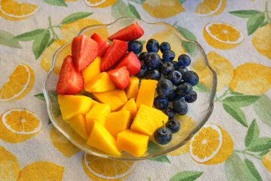 Glasschale mit Mangostücken, Erdbeerscheiben und Heidelbeeren auf einer Wachstischdecke mit gelbem Zitronenmuster.