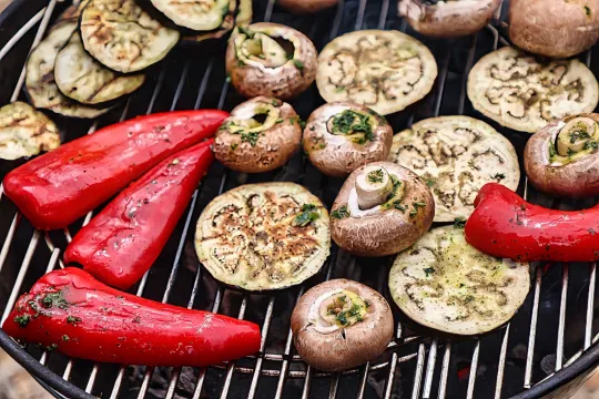 Draufsicht auf einen runden Grillrost, auf dem rote Spitzpaprika, Aubergienscheiben und mit Kräutern gefüllte Champignons über glühender Holzkohle brutzeln.