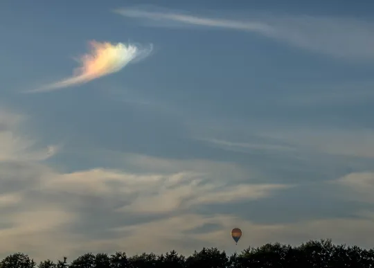 Leicht bewölkter blassblauer Himmel, an dessen unterem Rand eine dunkle Baumgrenze verläuft. Über den Bäumen schwebt klein ein Heißluftballon. Oben links leuchtet eine einzelne, federförmige Wolke in intensiven Regenbogenfarben.