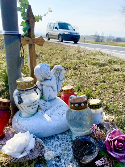 Provisorische Gedenkstätte am Straßenrand für ein Unfallopfer. Ein hölzernes Kreuz, ein schlafender Engel aus Stein auf einem herzförmigen Sockel mit der Aufschrift „Wir vermissen Dich“. Umgeben von mehreren Grablichtern in Rot und Weiß sowie künstlichen Blumen.