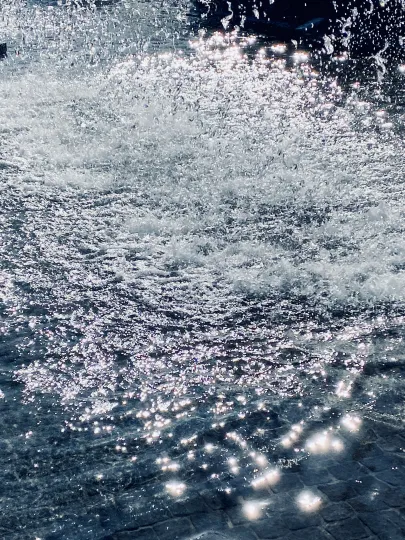  Sprudelnder Springbrunnen in hellem Sonnenlicht. Tausende glitzernde Wassertropfen spritzen über eine unruhige, blau schimmernde Wasseroberfläche, während das Licht in hellen, weißen Reflexionen auf dem Wasser tanzt. 