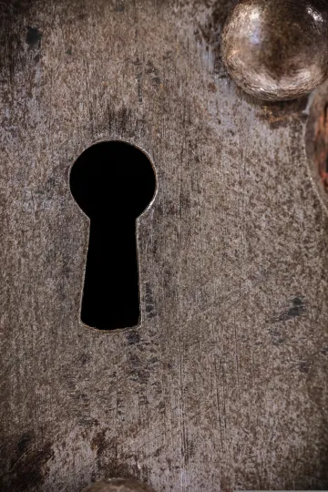 Schlüsselloch in einer massiven, metallischen Oberfläche. Das Metall wirkt alt, grob gebürstet und weist eine Textur aus Kratzern, Patina und leichten Korrosionsspuren auf.