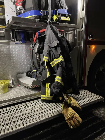 Eine Feuerwehrjacke mit gelben Reflektorstreifen hängt zusammen mit benutzten Lederhandschuhen an der geöffneten Geräteklappe eines Feuerwehrfahrzeugs in der Nacht.