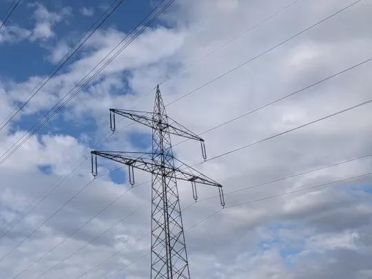 Ein großer Strommast aus Metall mit mehreren diagonal verlaufenden Stromleitungen vor einem teils blauen, teils bewölkten Himmel.