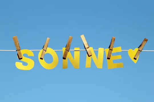 Aus gelbem Papier ausgeschnittenen Buchstaben des Wortes "SONNE" und ein kleines gelbes Herz hängen mit hölzernen Wäscheklammern an einer weißen Wäscheleine vor einem blauen, wolkenlosen Himmel