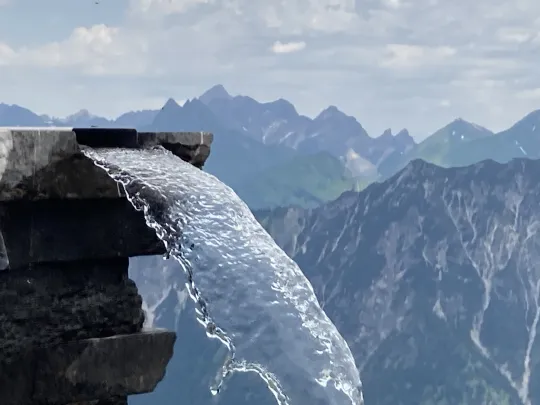 Im Bildvordergrund fließt aus einer steinernen Rinne ein breiter Wasserstrahl vor einem Panorama aus hohen Berggipfeln.