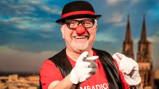 Lachender Clown mit roter Nase, Hut und weißen Handschuhen vor einer Luftaufnahme vom Kölner Dom