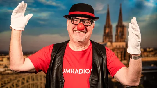 Lachender Clown mit roter Nase, Hut und weißen Handschuhen vor einer Luftaufnahme vom Kölner Dom