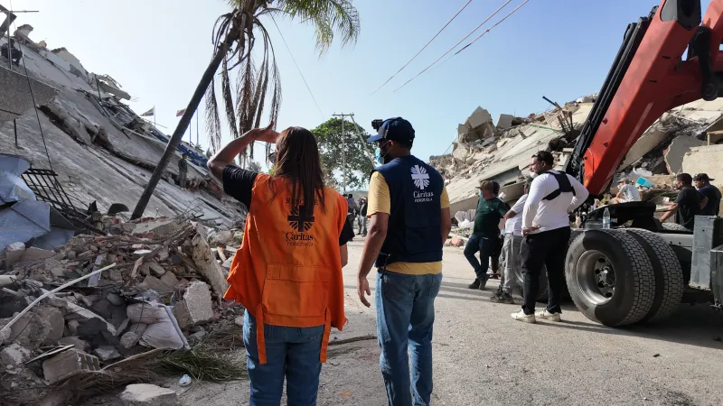 Zwei Mitarbeiter von Caritas International stehen auf de Straße vor einem von einem Erdbeben zerstörten Haus.