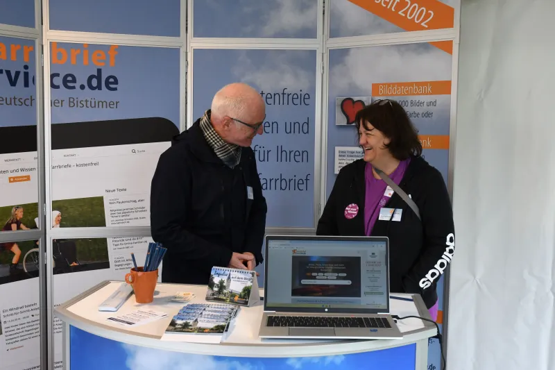 Zwei Personen unterhalten sich am Messestand von Pfarrbriefservice.de auf dem Katholikentag mit Laptop und Infomaterial.