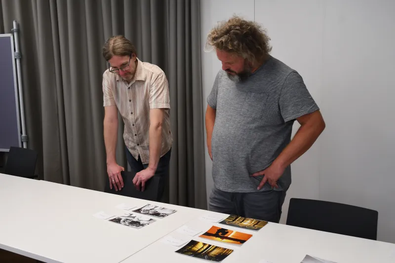 Zwei Männer betrachten Fotos, die auf einem weißen Tisch liegen.