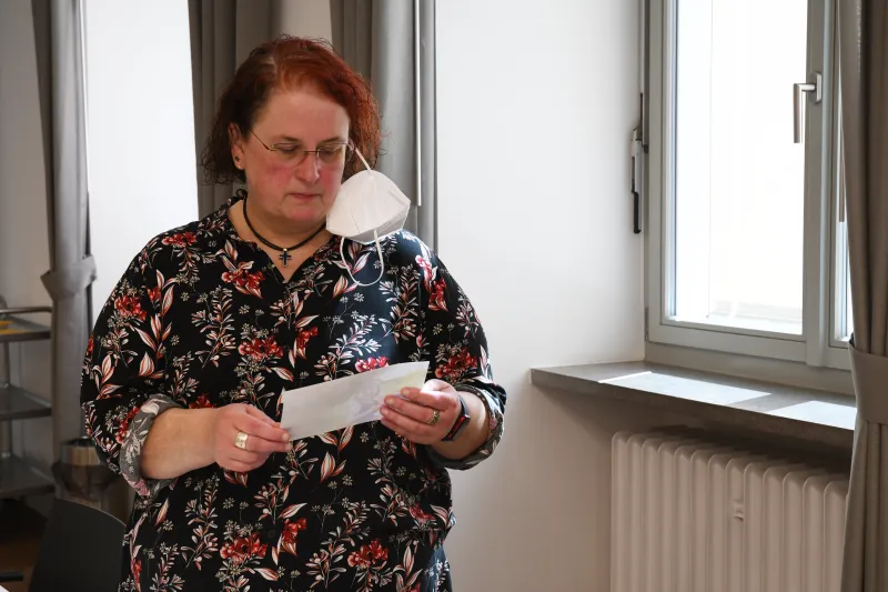 Frau mit Brille und Blumenbluse liest einen Zettel neben einem Fenster.