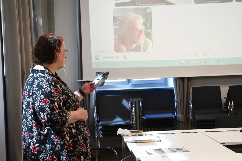 Frau mit Brille hält ein Foto vor einer Projektionsfläche in einem Besprechungsraum.