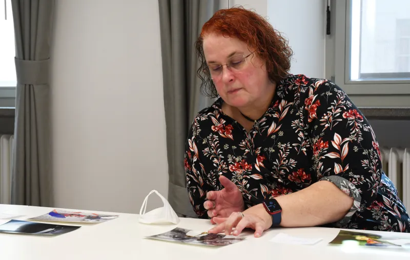 Frau mit rotem Haar betrachtet Fotos an einem Tisch, eine Maske liegt daneben.