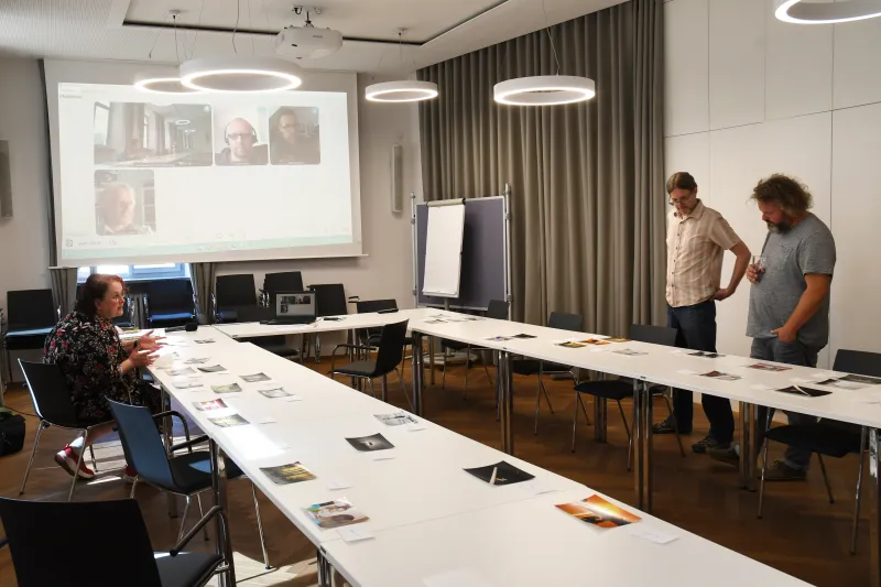 Heller Konferenzraum mit drei Personen, U-förmig gestellten Tischen und Projektion an der Wand.