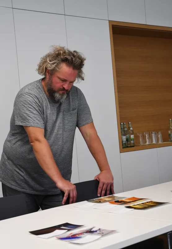 Mann mit grauem Shirt steht nachdenklich an einem Tisch mit Broschüren und Papieren.
