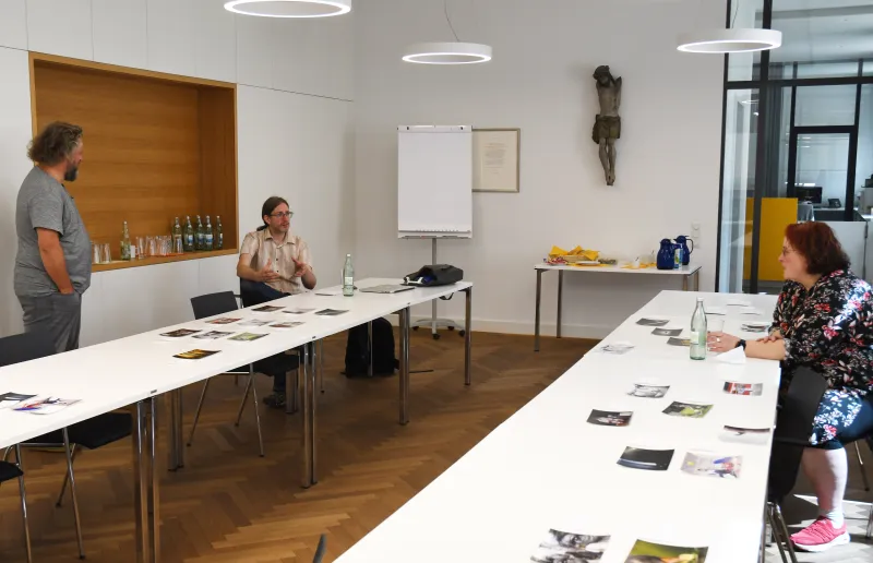 Drei Personen sitzen in einem Besprechungsraum mit Fotos auf weißen Tischen.