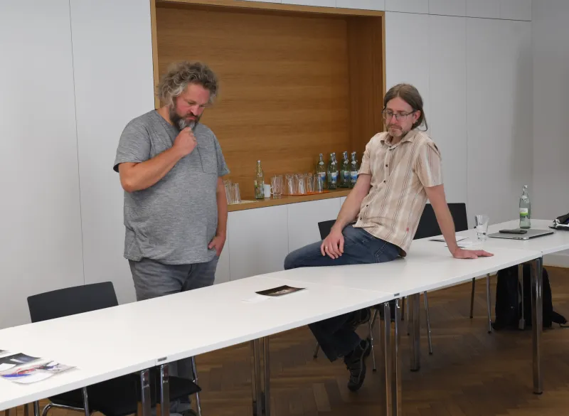Zwei Männer stehen nachdenklich an einem langen, weißen Konferenztisch.