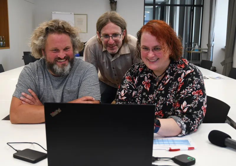 Drei lachende Personen sitzen gemeinsam vor einem Laptop in einem Besprechungsraum.