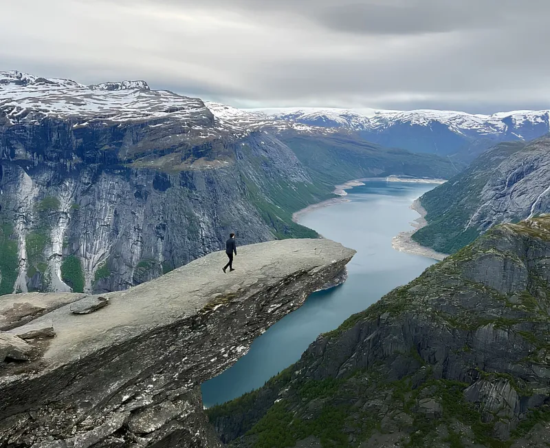 Person steht auf Felsvorsprung über Fjord mit Bergen und bewölktem Himmel
